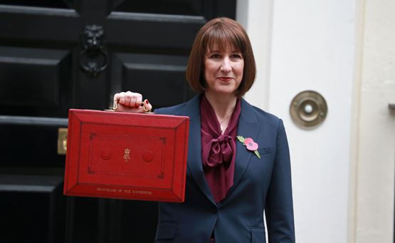 Chancellor of the Exchequer Rachel Reeves in a dark suit holding up a traditional red Budget box outside a black door, with a poppy pinned to her lapel