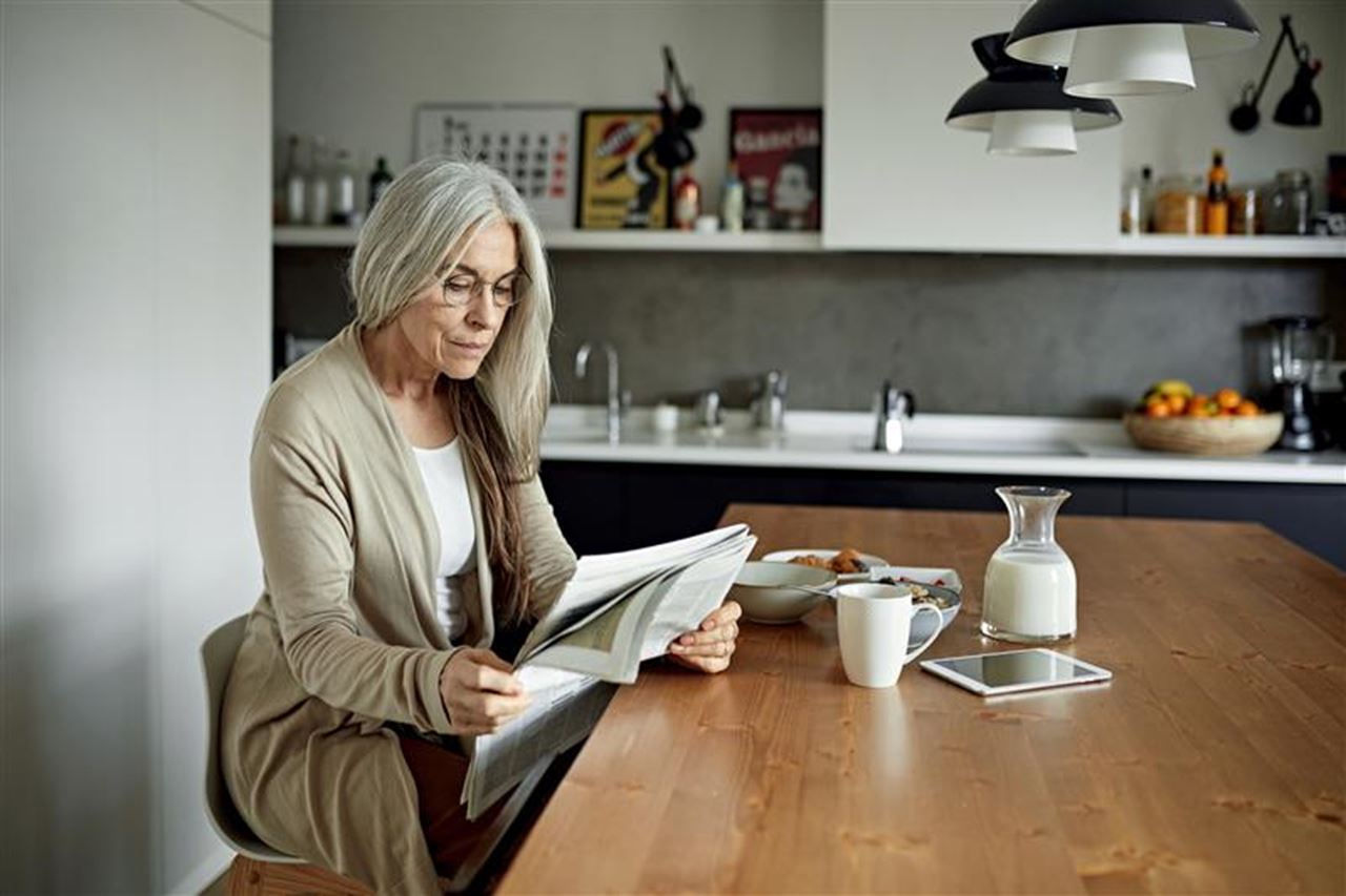 A woman sits at a dining table with breakfast in front of her, she is reading a newspaper.